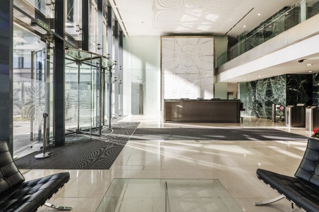 Atrium of an office building with green marble tiled walls, cream marble floors and a reception desk next to a revolving door at the entrance
