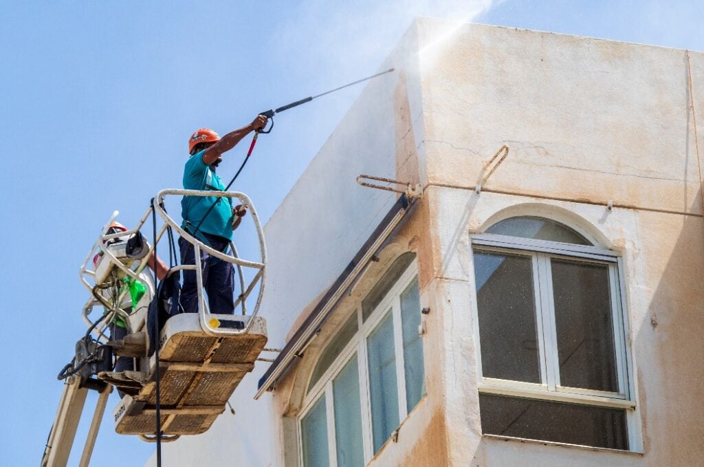 Hombre en una grúa realizando trabajos de limpieza de fachada con agua a presión en un edificio alto.